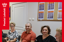 Na zdjęciu widać ludzi siedzących przy stole z dokumentami i wodą. W tle jest tablica z karteczkami oraz drzwi. Osoby patrzą w stronę aparatu, ubrane są elegancko.
