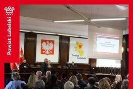 Sala wyłożona ciemnym drewnem, uczestnicy siedzą tyłem na krzesłach i słuchają mężczyzny stojącego przy stole prezydialnym, na ścianie godło Polski i mapa z napisem Powiat Lubelski, na ekranie slajd Zagrożenie z powietrza, szkolenie, Starostwo Powiatowe Lublin, 7 października 2026 rok. Grafika w czerwonej ramce, po lewej pionowy biały napis Powiat Lubelski z logo, na dole biały napis na czerwonym tle: Szkolenie w Starostwie Powiatowym w Lublinie.