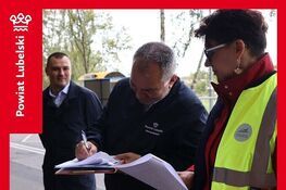 Trzy osoby stoją na zewnątrz w pobliżu drogi. Mężczyzna pośrodku podpisuje dokument. Kobieta w żółtej kamizelce patrzy na dokumenty, a inny mężczyzna stoi obok. W tle widoczne są drzewa.