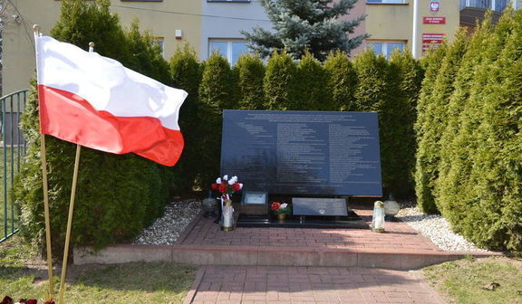 Pomnik w postaci płyty ozdobiony kwiatami i zniczami. Obok powiewają dwie flagi Polski. Całość otoczona szeregiem tui.