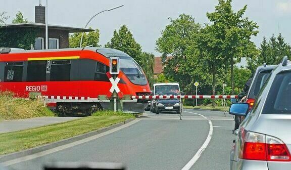 Czerwony pociąg przejeżdża przez przejazd kolejowy z opuszczonymi szlabanami, zatrzymując ruch samochodowy na drodze.