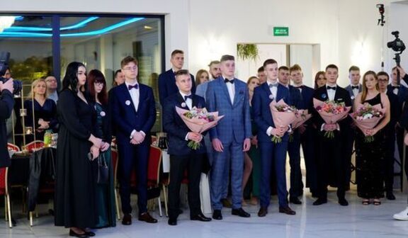 Grupa ludzi w eleganckim stroju stoi z bukietami kwiatów podczas oficjalnego wydarzenia w jasnym pomieszczeniu z kamerą w tle.