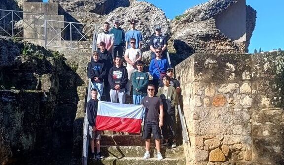 Grupa osób stoi na kamiennych schodach przed ruinami, trzymając polską flagę. Jasne niebo i zieleń w tle.