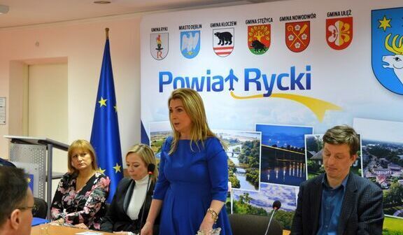 Na zdjęciu znajduje się grupa osób w biurowym wnętrzu. Kobieta w niebieskiej sukience stoi na tle plakatu z napisem "Powiat Rycki" oraz flagami Unii Europejskiej i Polski.