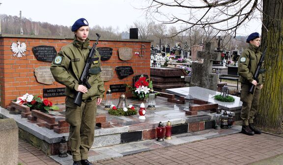 Dwóch żołnierzy w mundurach stoi na straży obok ceglanego pomnika z kwiatami na cmentarzu.