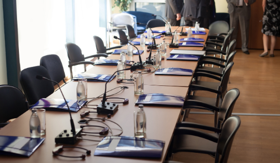 Zdjęcie przedstawia konferencyjny stół z ustawionymi krzesłami, mikrofonami oraz dokumentami, przygotowane na spotkanie w jasnym pomieszczeniu.