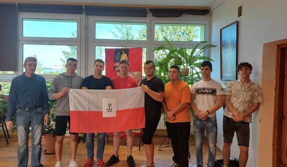 Ośmiu młodych mężczyzn stoi w rzędzie w pomieszczeniu z dużymi oknami, trzymają flagę Polski z godłem, po lewej stronie znajduje się zielona roślina.