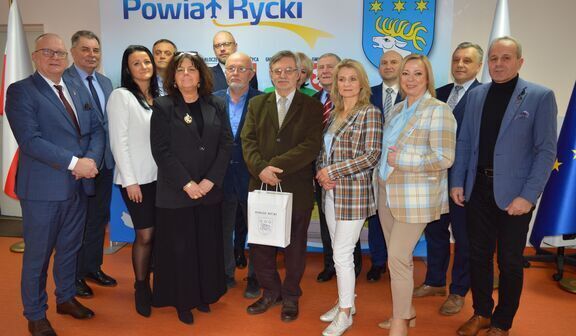Grupa ludzi uśmiecha się do kamery, stojąc w urzędowym pomieszczeniu z flagami Polski, Unii Europejskiej i emblematem "Powiat Rycki".