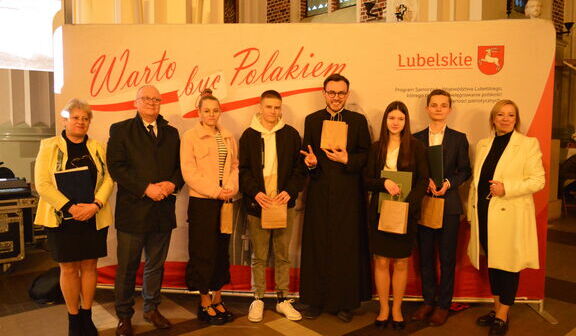 Grupa ośmiu osób stoi w rzędzie, trzymając certyfikaty. W tle widnieje napis "Warto być Polakiem" i herb Lubelskiego. Ubrania są eleganckie, nastrój uroczysty.