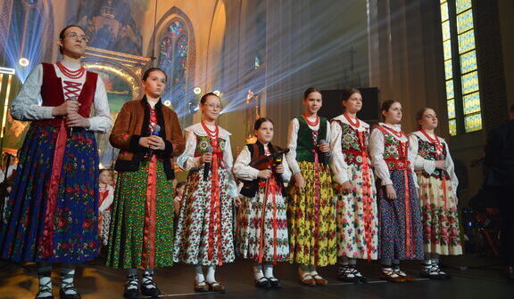 Grupa osób w tradycyjnych, polskich strojach ludowych stoi na scenie, z wnętrzem kościoła jako tłem.