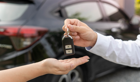 Osoba przekazuje klucze samochodowe do ręki drugiej osoby na tle niebieskiego samochodu.