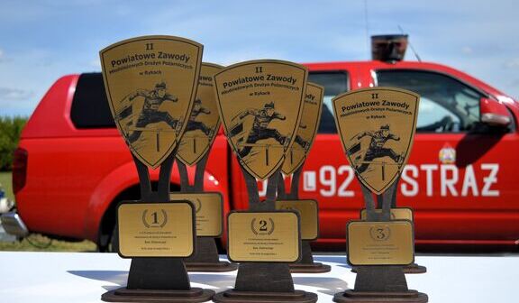 Trzy trofea z figurkami strażaków na pierwszym planie. Z tyłu czerwony wóz strażacki i niebieskie niebo.