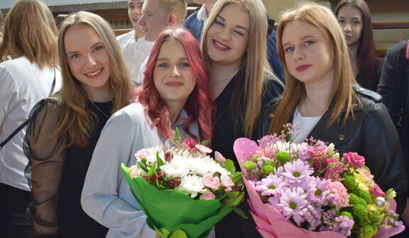 Cztery uśmiechnięte młode kobiety stoją obok siebie, trzymając bukiety kwiatów, w tle widoczni inni ludzie w dziennym świetle.