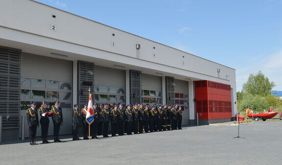 Grupa strażaków w ceremonialnych uniformach stoi przed remizą z otwartymi boksami, w których widać wóz strażacki; niebo jest pogodne.
