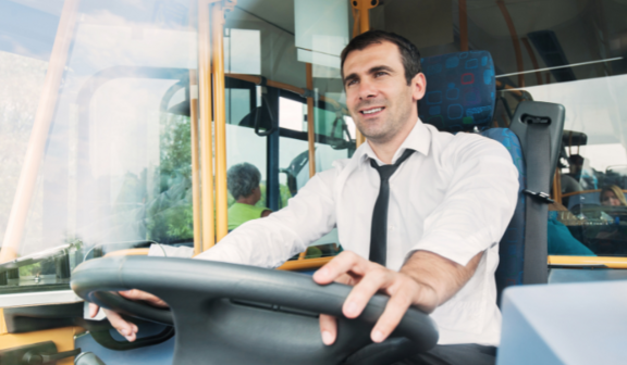 Kierowca autobusu w białej koszuli i krawacie trzyma kierownicę, uśmiechając się lekko. Siedzi on za panel sterowania pojazdu, w tle widoczni pasażerowie.