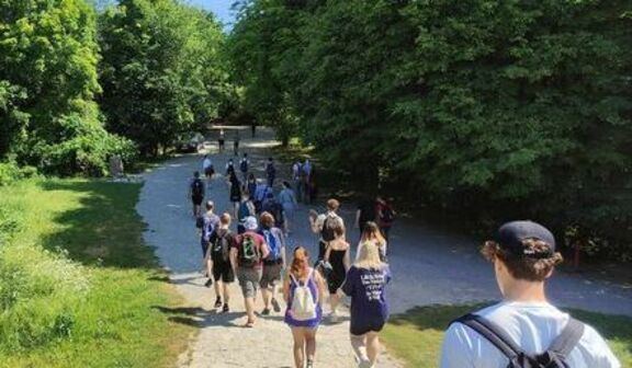Grupa ludzi spaceruje wzdłuż ścieżki w parku w słoneczny dzień, otoczona zielenią i drzewami.