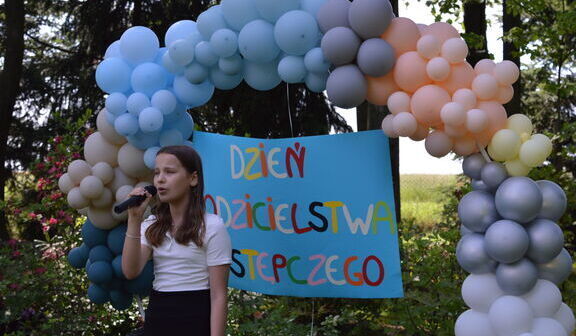 Dziewczyna śpiewa w mikrofon na tle kolorowej girlandy balonów i banera z napisem "Dzień Rodzicielstwa Zastępczego".