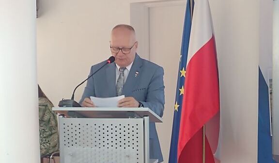 Mężczyzna w garniturze stoi za mównicą z logotypem "Powiat Rycki" i flagami Polski oraz Unii Europejskiej w tle, czyta dokument.