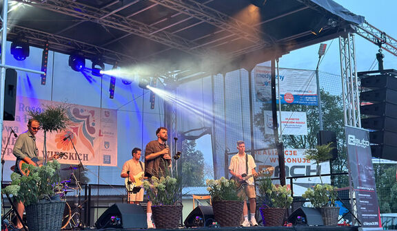Na scenie podczas koncertu na świeżym powietrzu stoi czteroosobowy zespół; artyści grają na gitarach i śpiewają, otoczeni sprzętem muzycznym i roślinami w doniczkach.