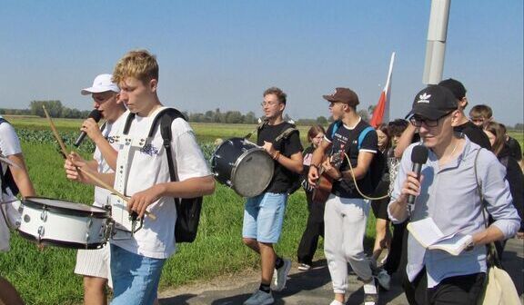 Grupa ludzi maszeruje wzdłuż drogi podczas dnia o słonecznej pogodzie; niektórzy grają na instrumentach takich jak bęben i flet.