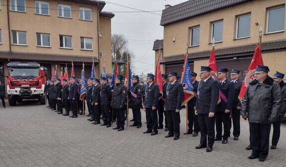 Strażacy w mundurach ustawieni w linię z flagami przed budynkiem i wozem strażackim.