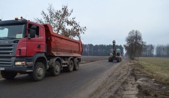 Ciężarówka z czerwoną przyczepą stoi na wiejskiej drodze, a z przodu widać koparkę pracującą przy poboczu. Wokół znajduje się otwarta przestrzeń z drzewami i polami.