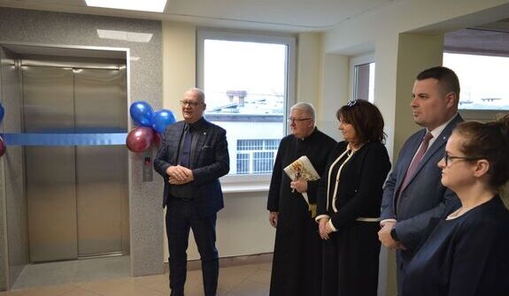 Grupa osób przy windzie podczas uroczystości otwarcia. W tle widać balony. Osoba przemawiająca stoi obok włącznika windy. Reszta osób obserwuje i trzyma dokumenty oraz kwiaty.