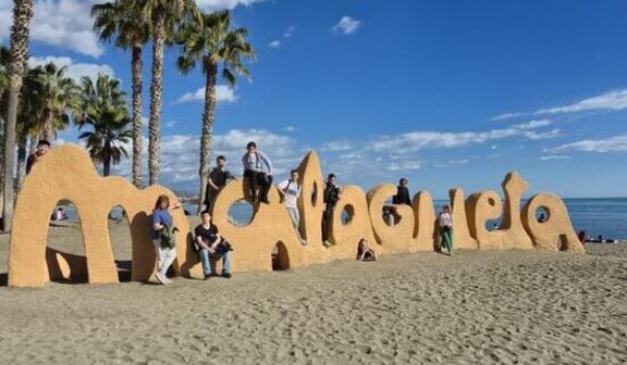 Grupa osób pozuje przy dużym, piaskowym napisie "Malagueta" na plaży. W tle widać palmowe drzewa, morze i częściowo zachmurzone niebo. Ludzie wspinają się na litery, ciesząc się pogodnym dniem.