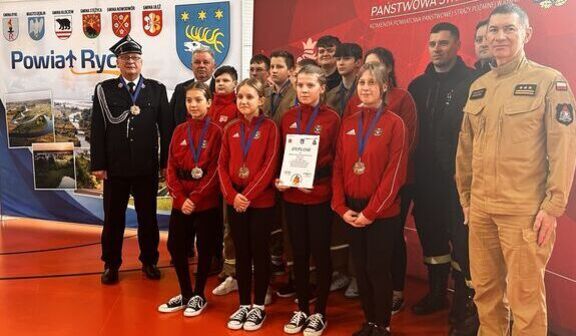 Grupa osób na podium podczas wydarzenia związanego ze strażą pożarną. Osoby są ubrane w czerwone bluzy z medalami i trzymają dyplom. W tle widoczne są banery z emblemami i napisami.