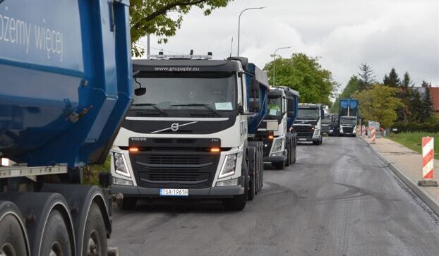 Rząd ciężarówek z przyczepami w kolorze niebieskim jedzie po świeżo wyasfaltowanej drodze. W tle drzewa i część zabudowy mieszkalnej. Na poboczu widoczne są pachołki ostrzegawcze.