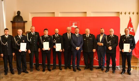Grupa mężczyzn stoi w dwóch rzędach w formalnych strojach. Kilku z nich trzyma dokumenty. W tle czerwone tło z logotypem i polska flaga. W rogu znajduje się popiersie.