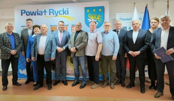Grupa osób stoi przed banerem z napisem "Powiat Rycki". W tle herb i flagi. Większość osób jest elegancko ubrana. Mają poważne wyrazy twarzy, a niektórzy trzymają dokumenty.