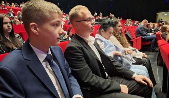Grupa ludzi siedząca w teatrze na czerwonych fotelach. Wszyscy elegancko ubrani, głównie w garnitury i formalne stroje. Publiczność w tle również skupiona na wydarzeniu na scenie.