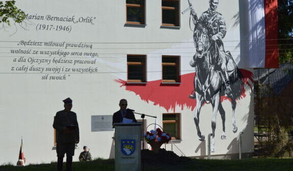 Na ścianie budynku mural z żołnierzem na koniu oraz cytatem. Obok podium z mężczyzną przemawiającym oraz żołnierzem w mundurze. W tle cień drzewa daje kontrastowy efekt na fasadzie.
