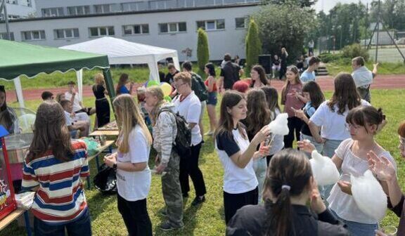 Grupa ludzi na szkolnym festynie na zewnątrz. Uczestnicy stoją pod namiotami, trzymają watę cukrową i rozmawiają. W tle budynek szkoły i drzewa. Atmosfera jest swobodna i wesoła.