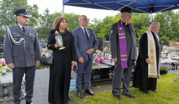 Grupa osób uczestniczy w ceremonii na cmentarzu. Dwie osoby w mundurach wojskowych, jedna trzyma lampion. Dwóch duchownych w szatach liturgicznych. Stoją pod niebieskim namiotem, wokół są groby i kwiaty.