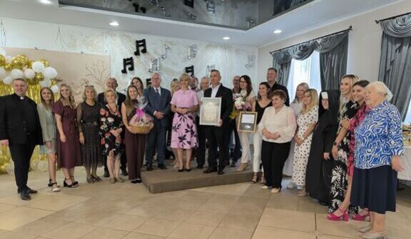 Grupa osób pozująca podczas uroczystości w eleganckim wnętrzu, z dekoracjami z balonów i nut. Niektórzy trzymają dyplomy i kosze upominkowe. Wszyscy uśmiechnięci, ubrani odświętnie.