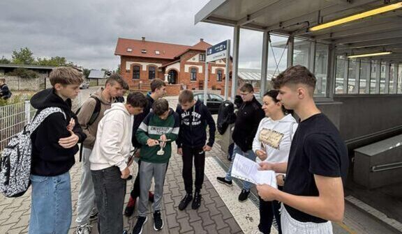 Grupa ludzi, w tym młodzież, stoi na peronie kolejowym. Dwójka z nich trzyma kartki. W tle ceglany budynek i tabliczka informacyjna. Niebo jest pochmurne.