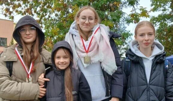 Cztery dziewczyny stoją na zewnątrz, ubrane w ciepłe kurtki i medale na szyi. W tle drzewa z jesiennymi liśćmi. Wszystkie uśmiechają się do zdjęcia.