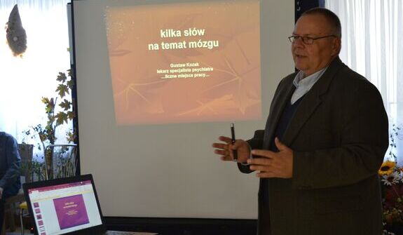 Mężczyzna w garniturze prowadzi prezentację na temat mózgu. Stoi obok ekranu z tytułem "kilka słów na temat mózgu". W tle laptop z otwartą prezentacją. Pomieszczenie jest jasne, z zasłoniętymi oknami.