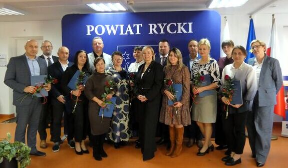 Grupa ludzi stoi uśmiechnięta, trzymając kwiaty i teczki. W tle znajduje się niebieska tablica z napisem "Powiat Rycki". Pomieszczenie jest jasne, a na ścianie wiszą flagi.