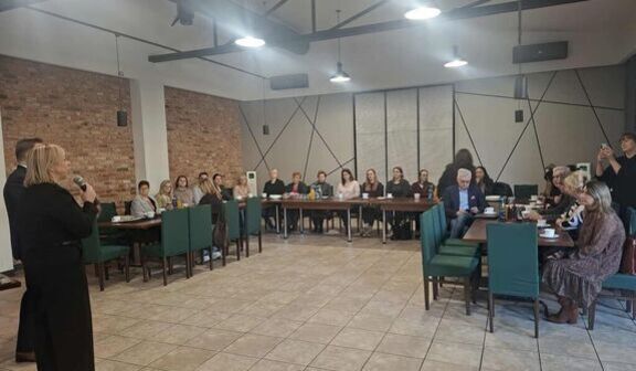 Grupa osób siedzi przy długich stołach w sali konferencyjnej z ceglanymi i szarymi ścianami. Kobieta z mikrofonem stoi z boku, reszta słucha, patrząc w jej kierunku. Na stołach leżą notatki i napoje.