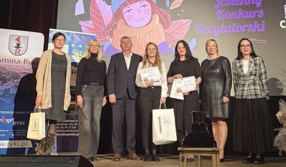 Grupa osób stoi na scenie z nagrodami i dyplomami, uśmiechają się do zdjęcia. W tle widać banery i kolorowy plakat wydarzenia podświetlany reflektorami.