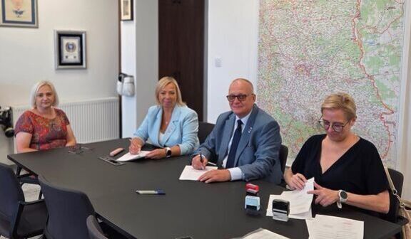 Cztery osoby siedzą przy stole w sali konferencyjnej, podpisując dokumenty. Na ścianie obok znajduje się duża mapa.