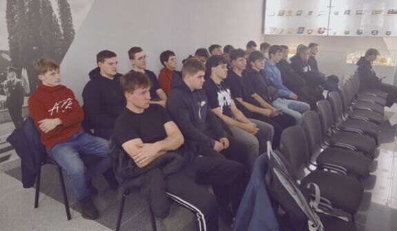 Grupa młodych ludzi siedzi w szeregu na krzesłach w nowoczesnym pomieszczeniu. Po lewej stronie jest czarno-biała grafika drzewa, a po prawej przeszklona szafka z różnymi przedmiotami na wystawie.