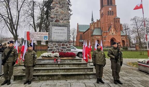 Grupa żołnierzy stoi na placu przed pomnikiem i kościołem z czerwonej cegły. Wokół pomnika znajdują się biało-czerwone flagi i kwiaty. W tle widoczny jest kościół i drzewa.