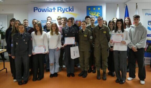 Na zdjęciu znajduje się grupa młodych ludzi stojących w dwóch rzędach. Niektórzy z nich trzymają dyplomy. W tle widać plakaty i flagi. Osoby są w różnym ubraniu, zarówno formalnym, jak i codziennym.