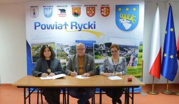 Na zdjęciu troje ludzi siedzi przy stole z dokumentami. W tle znajduje się baner z napisem „Powiat Rycki”, z herbami gmin i flagami Polski oraz Unii Europejskiej.