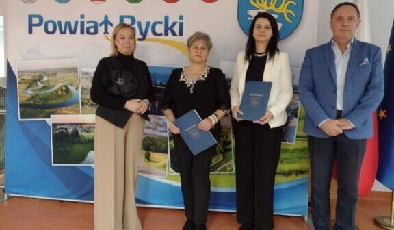 Cztery osoby stoją na tle ścianki z napisem "Powiat Rycki" i herbami gmin. Trzy kobiety i jeden mężczyzna trzymają dokumenty. Obok widoczne są flagi.