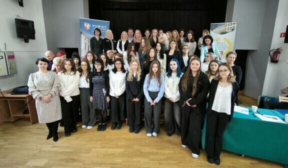 Grupa ludzi stoi na tle banerów. Osoby ubrane są elegancko. W tle znajduje się scena z ciemną kurtyną. Po prawej stronie jest stolik z mikrofonem. Całość odbywa się w przestrzeni zamkniętej.